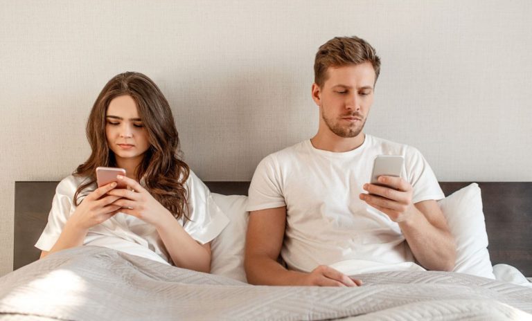 Couple in bed on their cell phones, symbolizing disengagement and the importance of connection in Strengths-Based Couples